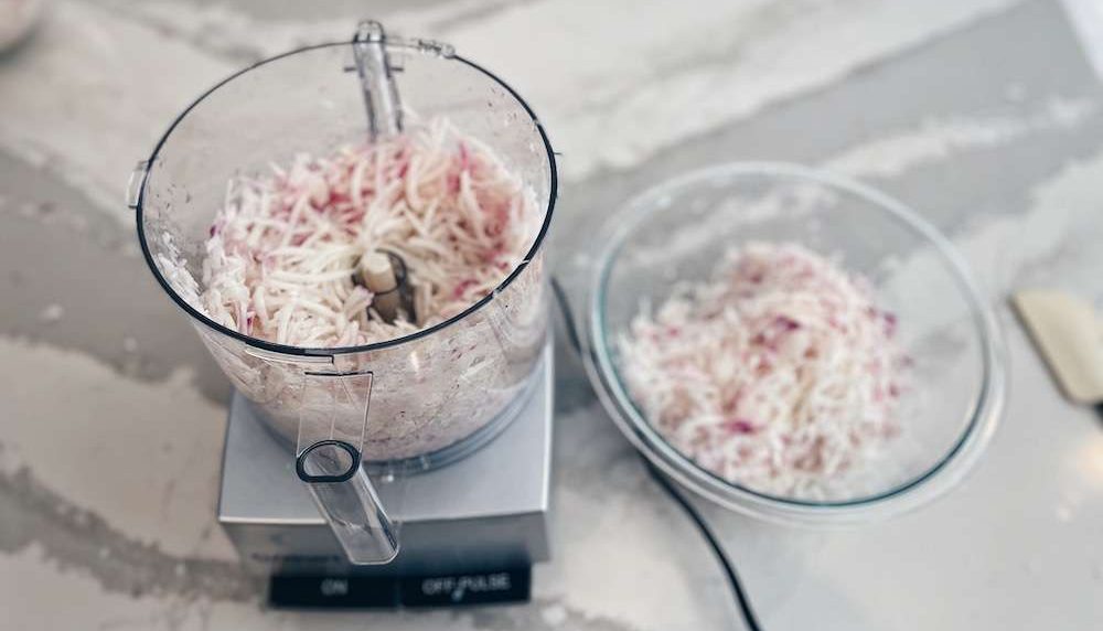 A food processor next to a glass bowl full of shredded turnips