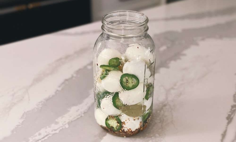 Hard-boiled eggs, jalapenos and spices added to a half gallon mason jar sitting on a kitchen island