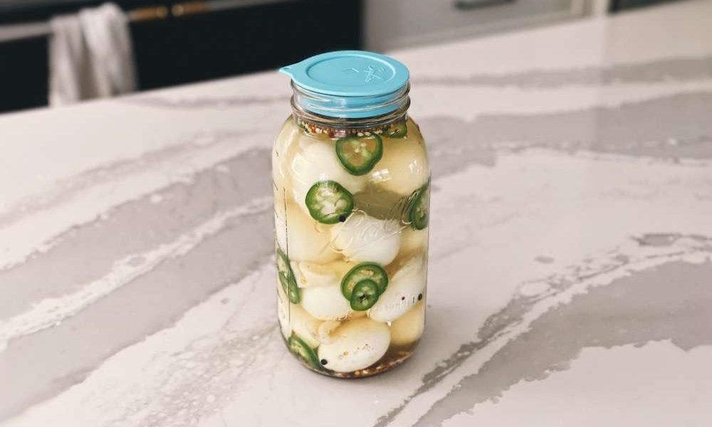 A jar of finished jalapeno pickled eggs sitting on a kitchen island with a blue silicone lid on top