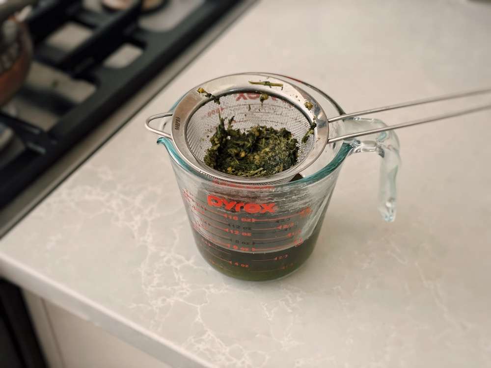 A glass measuring cup holding the strained out chickweed oil and a strainer full of chickweed herbs on top