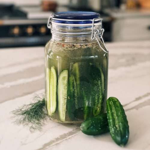A jar of refrigerator pickles sitting on the kitchen island with 2 cucumbers and fresh dill next to it.
