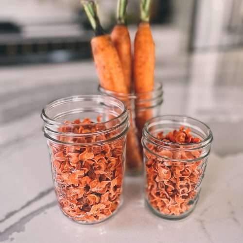 2 glass jars filled with dehydrated carrots in front of 3 whole carrots also in a mason jar
