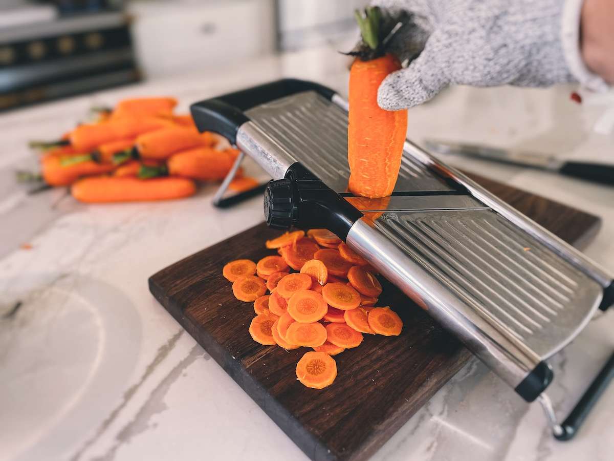 Slicing carrots using a mandoline slicer and wearing a cut-proof glove on top of a wooden cutting board.