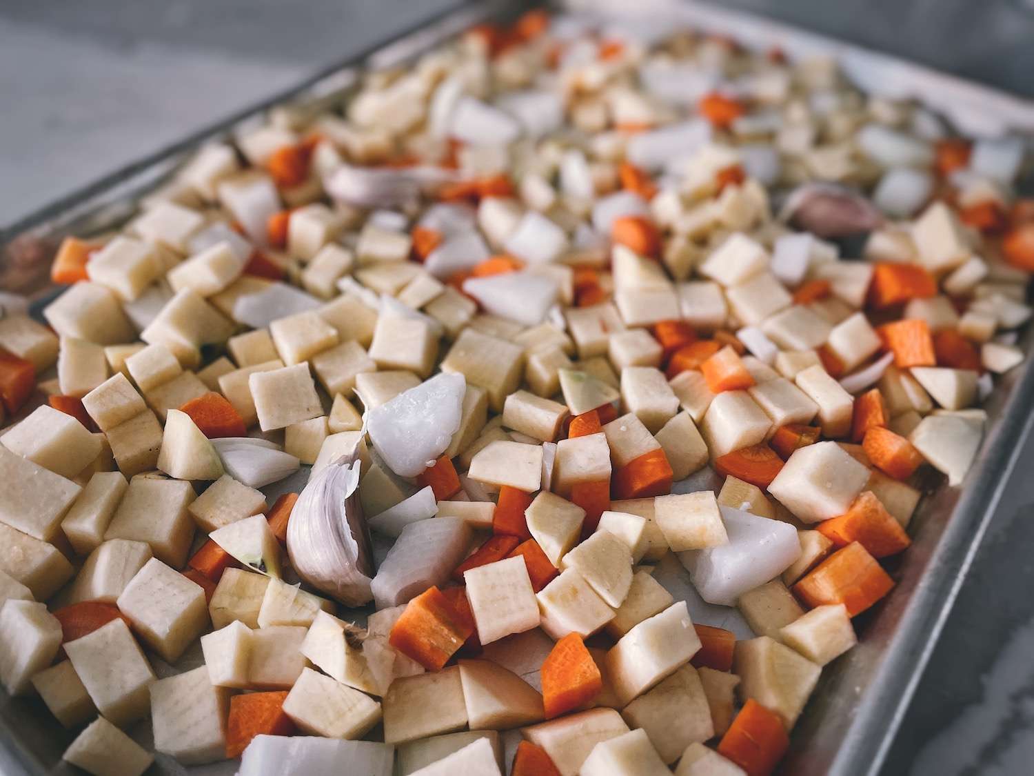 Chopped swede, onions, and carrots on a sheet pan along with garlic cloves and coconut oil.