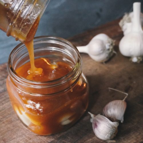 A photo of a jar full of raw garlic with raw honey being poured over it. The jar is sitting on a wooden cutting board on a slate countertop.