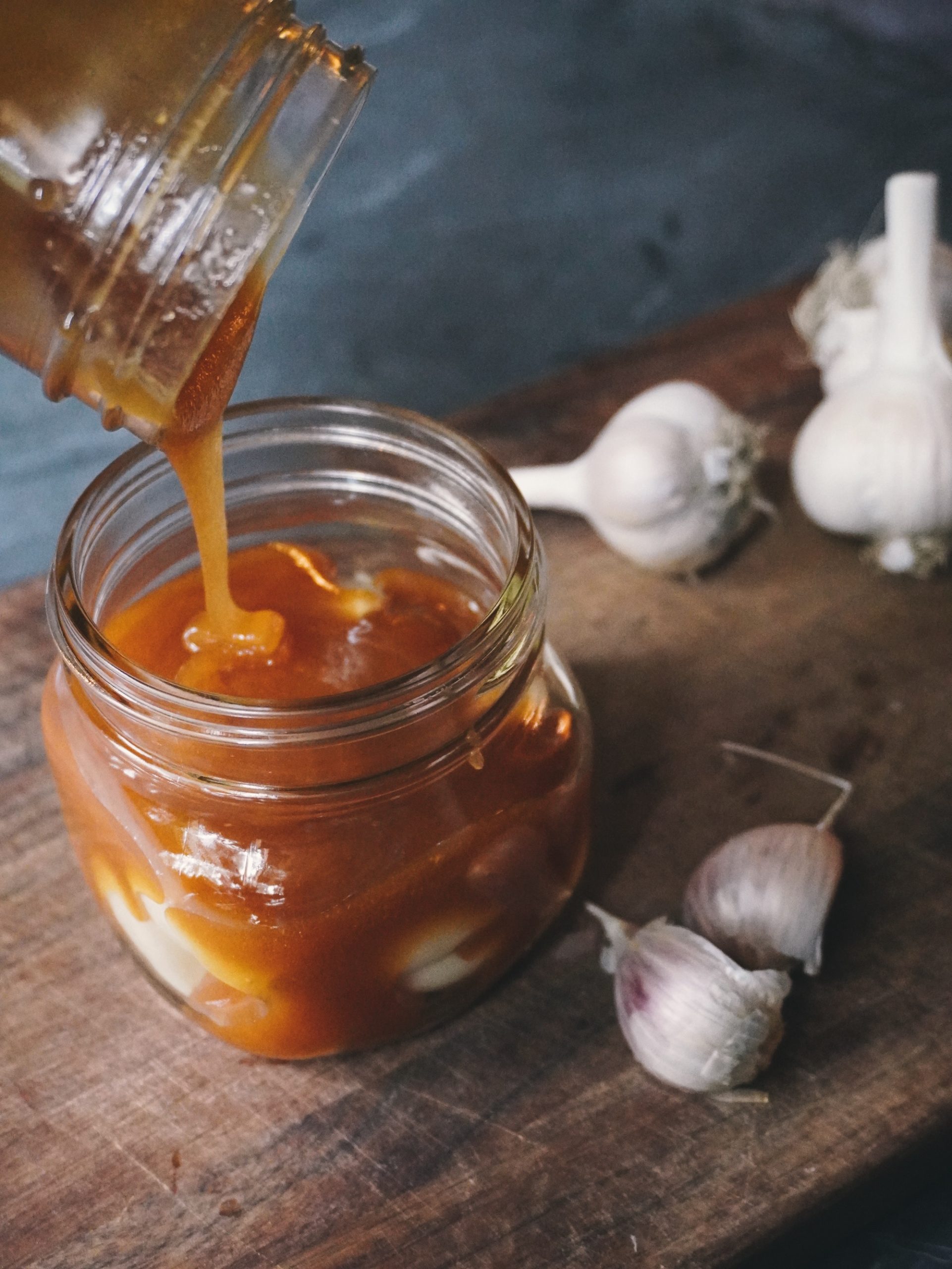 A photo of a jar full of raw garlic with raw honey being poured over it. The jar is sitting on a wooden cutting board on a slate countertop.