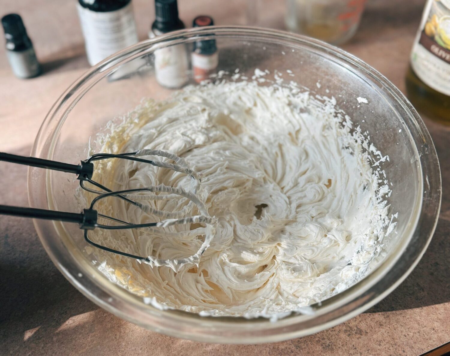 A photo showing a hand mixer whipping up tallow balm with essential oil bottles in the background.