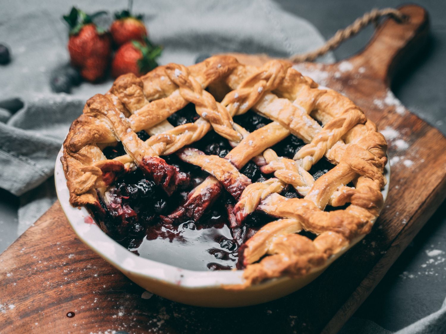 A blueberry pie with a cross hatch pattern sitting on a wooden background. Strawberries sitting on the side.