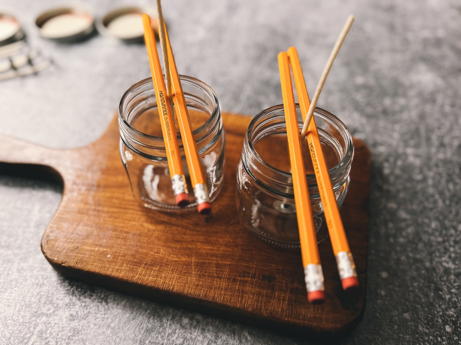 A photo showing candle wicks suspended in empty glass jars and being held in place with 2 pencils