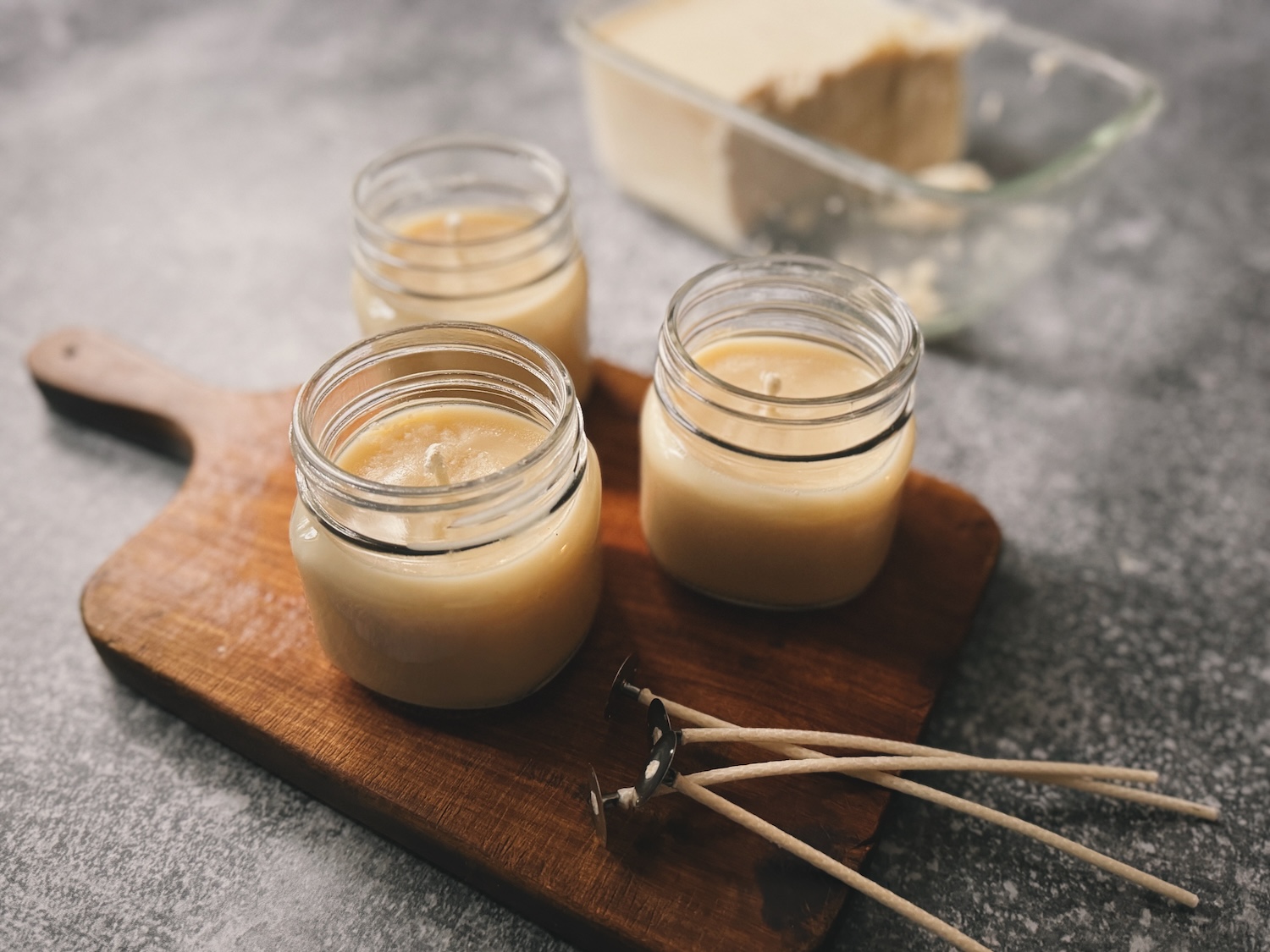 3 tallow candles in glass jars sitting on a wooden cutting board with unused wicks nearby