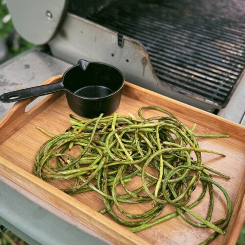 Grilled garlic scapes are resting on a wooden tray with a black cast iron small pot full of tallow near a grill