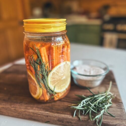 A quart mason jar filled with fermented carrot sticks. Sitting on a wooden cutting board.