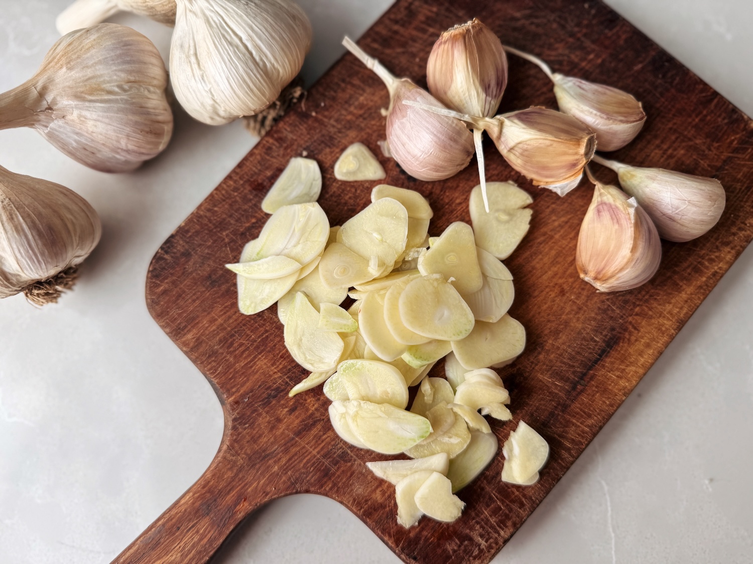 Thinly sliced garlic is piled up on a wooden cutting board with whole garlic nearby