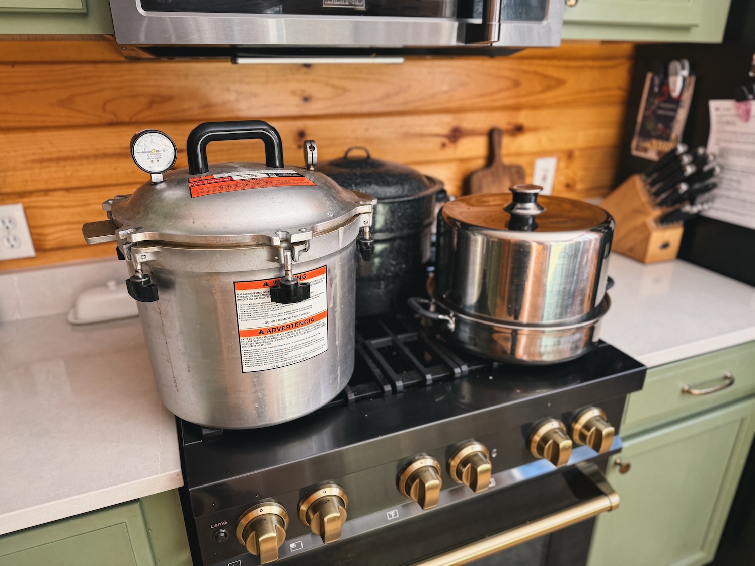 A pressure canner, water bath canner, and steam canner are sitting on a range in a kitchen. The photo is taken from the side